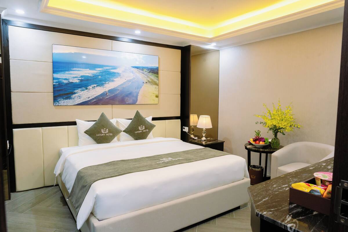 A hotel room with a large bed, a nightstand with a lamp, a chair, a desk with a vase of flowers, and a painting of a beach on the wall.