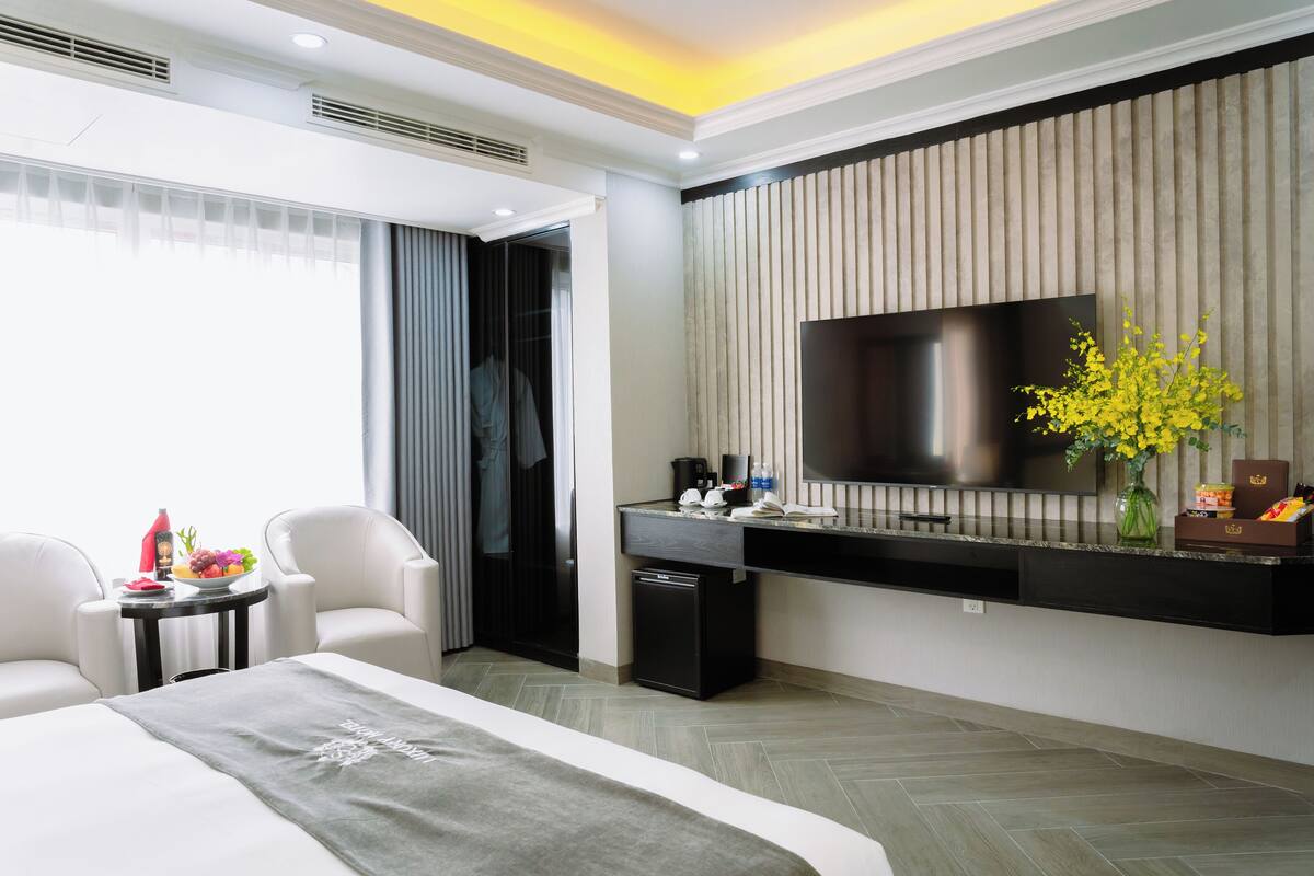 A modern hotel room with a large bed, a desk with a flat-screen TV, a small refrigerator, and a seating area with two chairs.