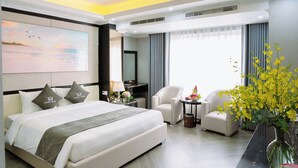 A modern hotel room with a large bed, a flat-screen TV, a desk with a lamp, two armchairs, and a vase with yellow flowers. - SC Luxury Hotel (Bai Chay)
