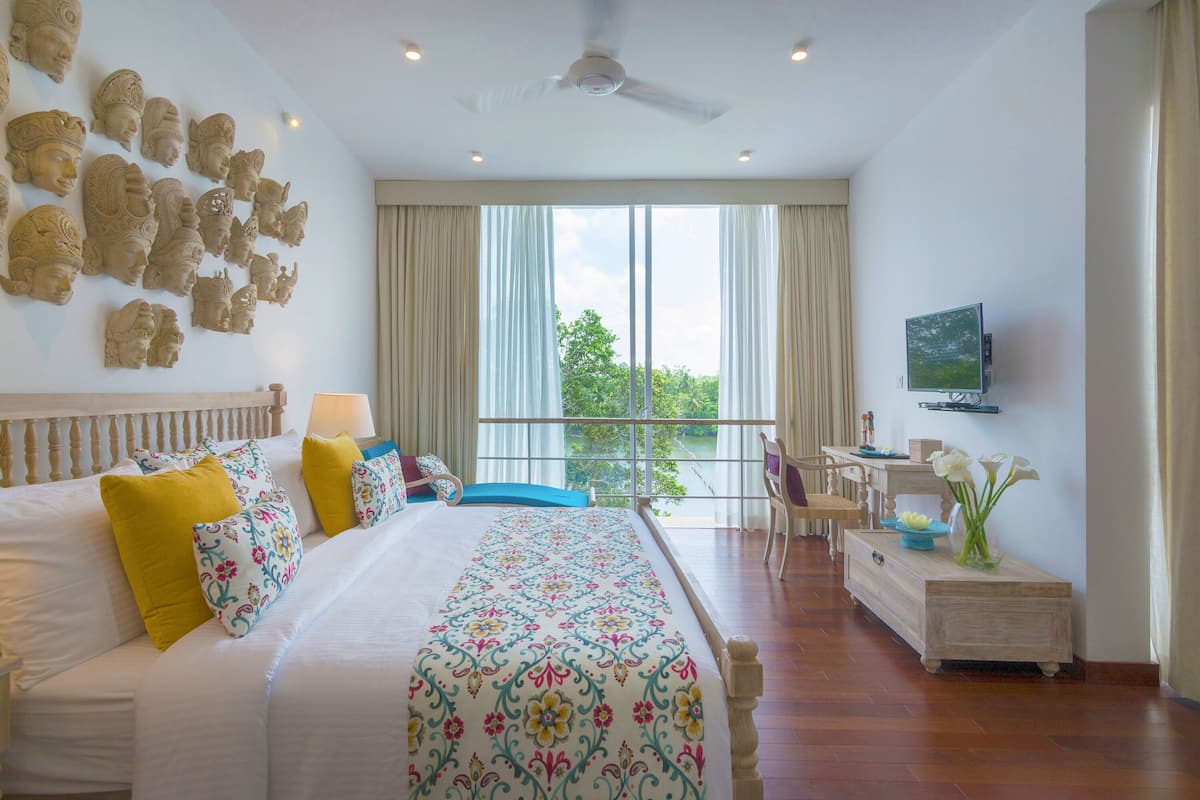 A hotel room with a large bed, a desk, a television, and a balcony with a view of the greenery outside.