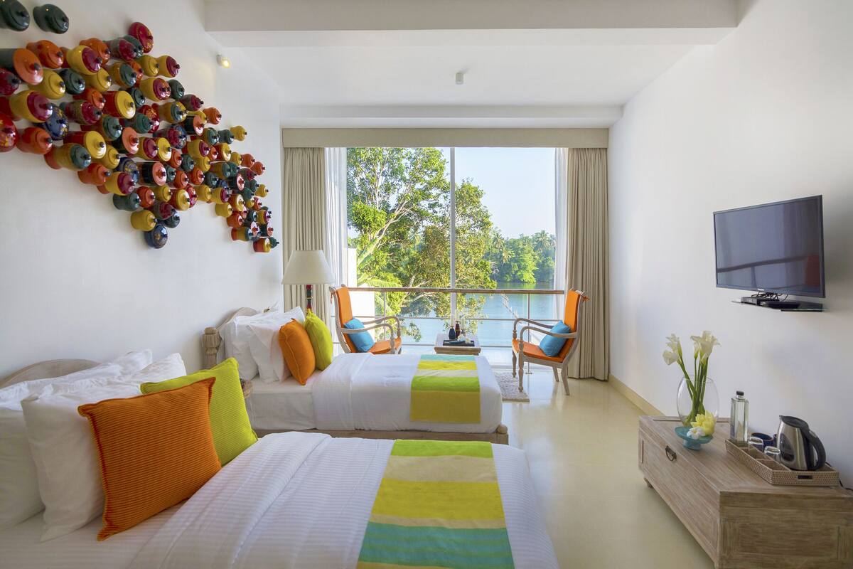 A hotel room with two beds, a TV, a balcony with a view of trees, and a wall art installation.