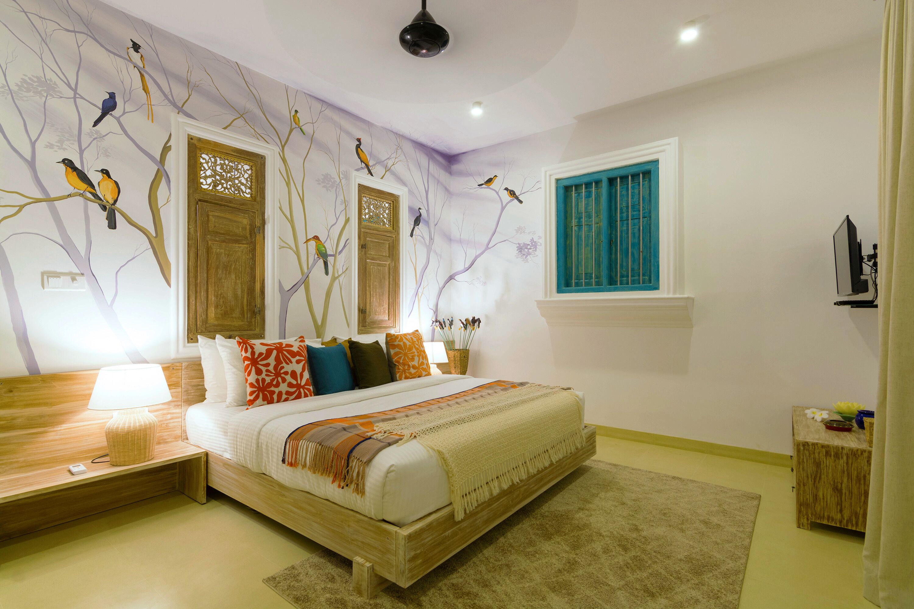 A bedroom with a bed, nightstands, a lamp, a window with blue shutters, and wall art of birds on trees.