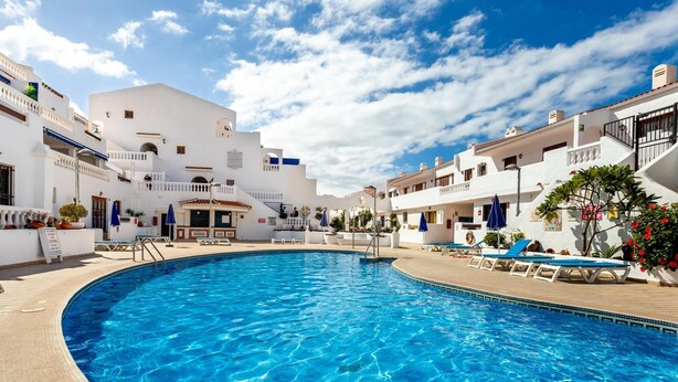 A swimming pool surrounded by white buildings with balconies, a clear sky, and a wooden deck. - Port Royale shine and bright, heated pool (Los Cristianos)