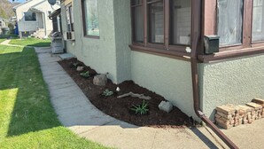 Una casa con jardín delantero, un sendero y una pequeña partera con plantas y rocas.