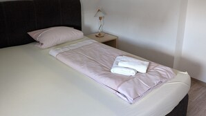 Een netjes opgemaakt bed met hoofdeinde, een nachtkastje met een lamp, en een effen muur.