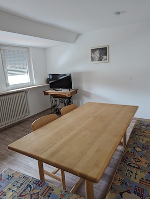 Una habitación con una mesa de madera, un televisor sobre un mueble bajo, un radiador, una ventana con persianas y una alfombra estampada.