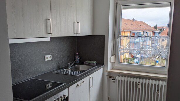 Een moderne keuken met een zwart aanrecht, witte kasten, een inbouwoven en een raam met uitzicht op gebouwen.