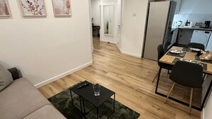 Ein modernes Wohnzimmer mit einer Couch, einem Couchtisch und einem Essbereich mit Tisch und Stühlen. Es gibt eine Küchenzeile mit einem Kühlschrank und einer Waschmaschine.