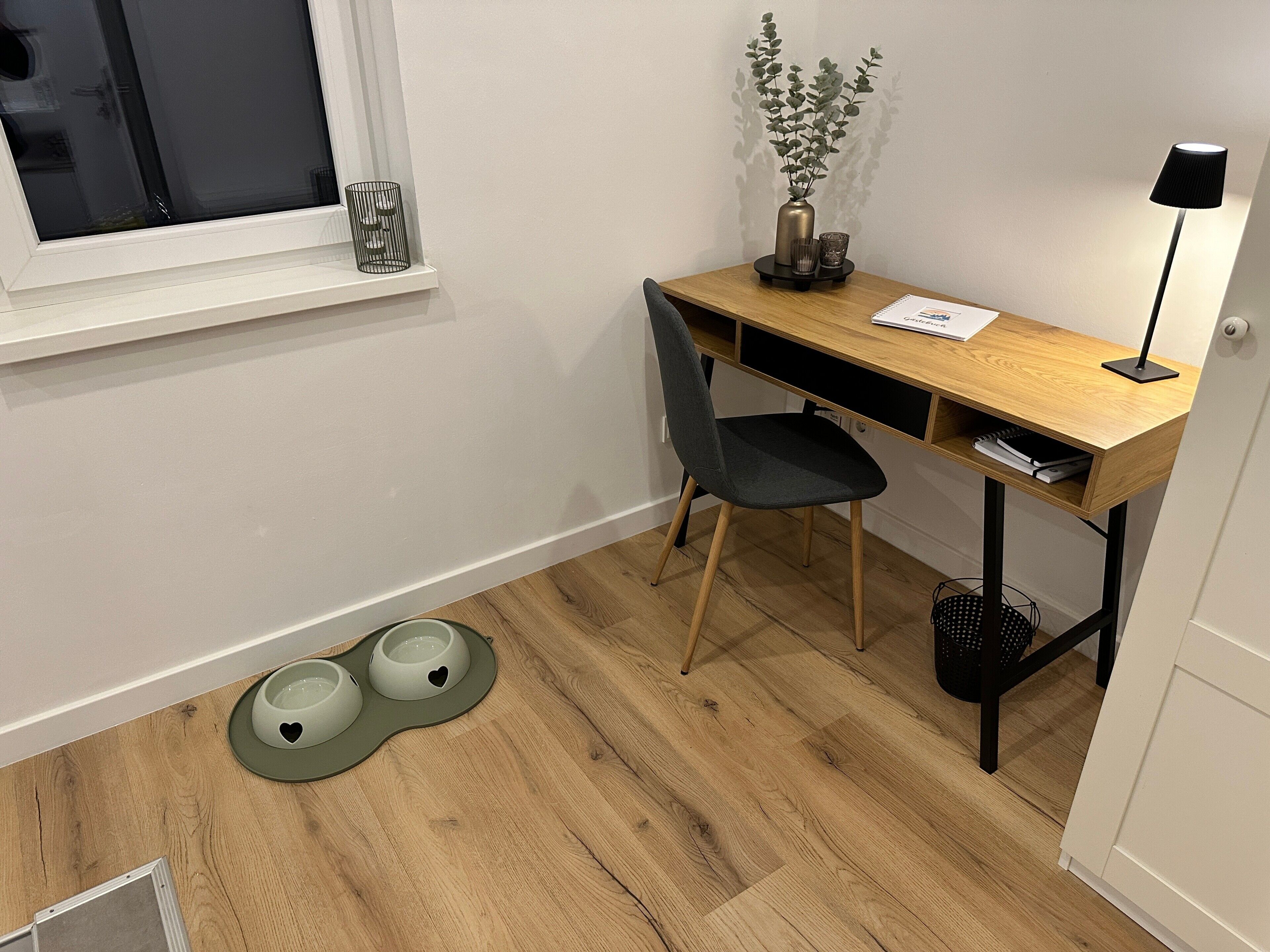 A room with a wooden desk, a chair, a window, a plant, a lamp, and a pet feeding station.