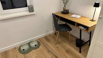 A room with a wooden desk, a chair, a window, a plant, a lamp, and a pet feeding station.