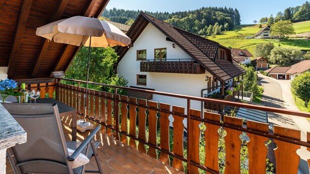Een balkon met een houten hekwerk, een tafel met stoelen en uitzicht op een wit huis met balkon.