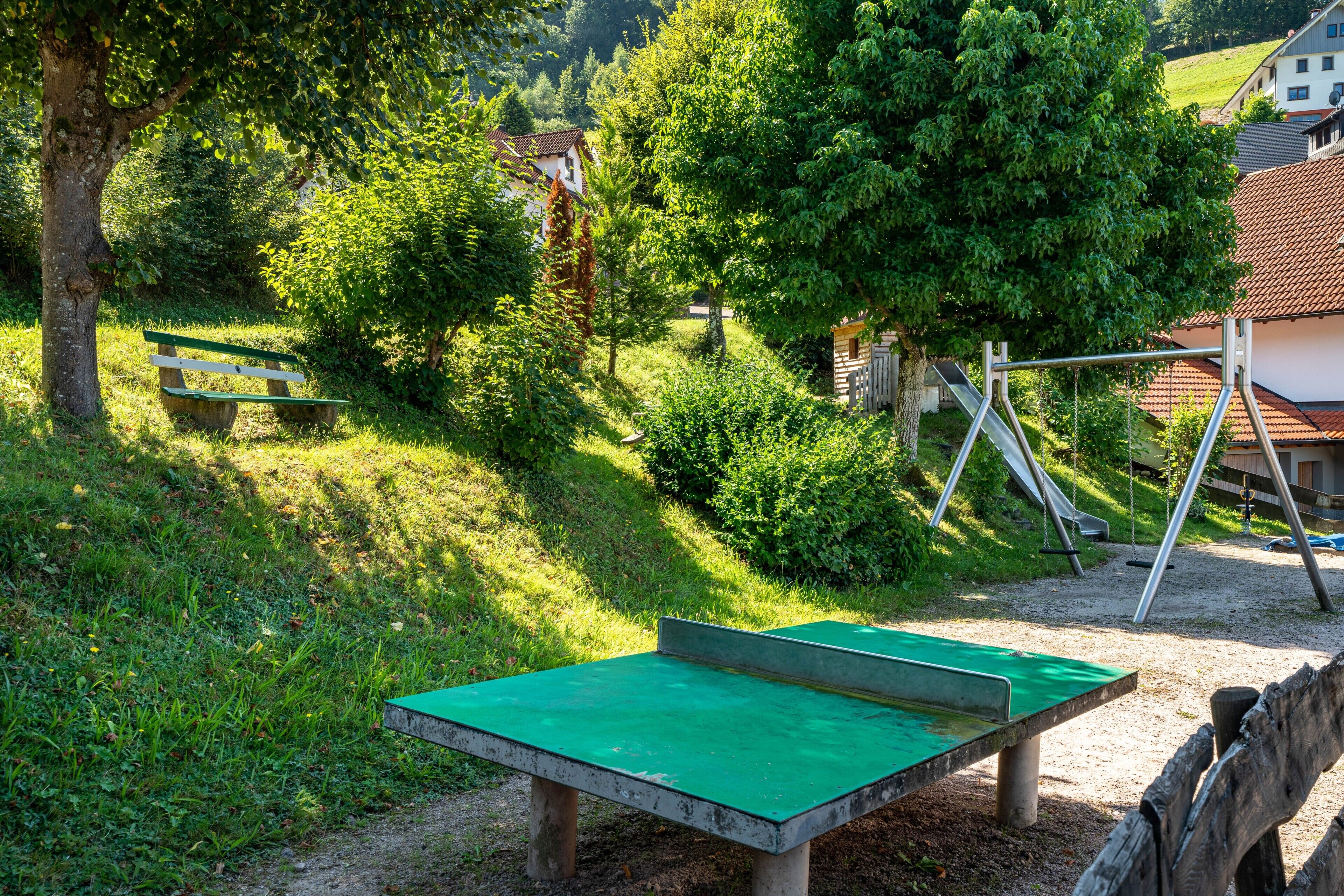 Eine grüne Parkbank, eine Schaukel und ein Spieltisch in einem Park mit Bäumen und Häusern im Hintergrund.