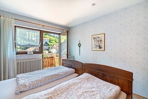 Une chambre avec deux lits, une fenêtre donnant sur une maison et sur de la verdure, un radiateur et un tableau encadré au mur.
