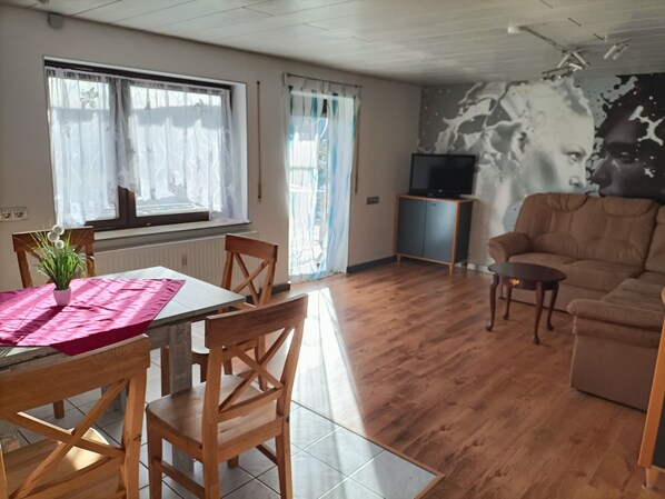 A living room with a wooden floor, a dining table with chairs, a sofa, a television, and a wall with a partial mural.