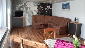 A living room with a sectional sofa, a small wooden table, a TV on a cabinet, and a wall mural. - Apartment 'Bamboleo' with Shared Terrace, Private Garden and Wi-Fi (Veringendorf)