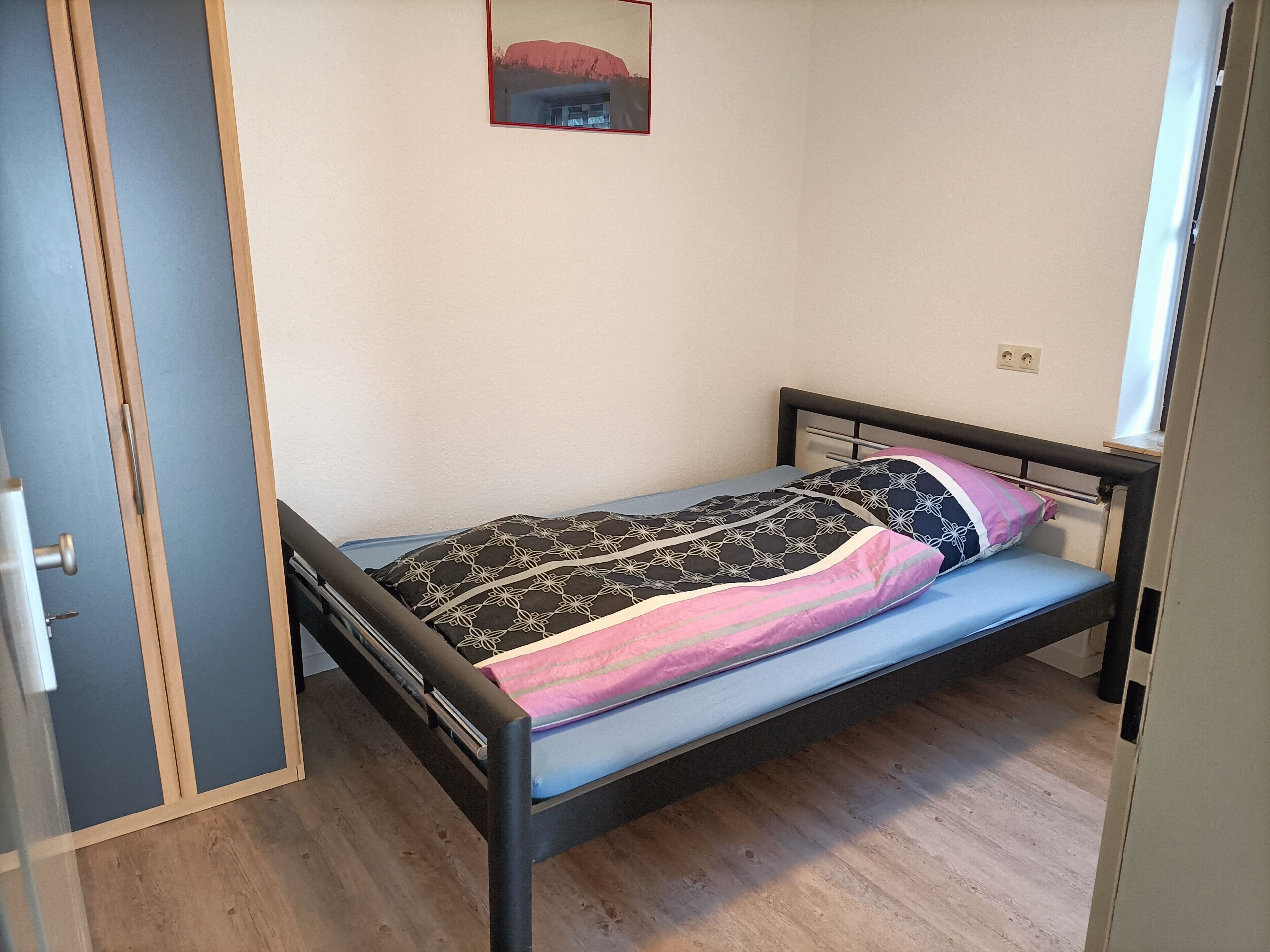 Ein Einzelbett mit einem gemusterten Bettbezug und Kissen, ein Holzboden, eine Tür und ein an der Wand hängendes Bild.