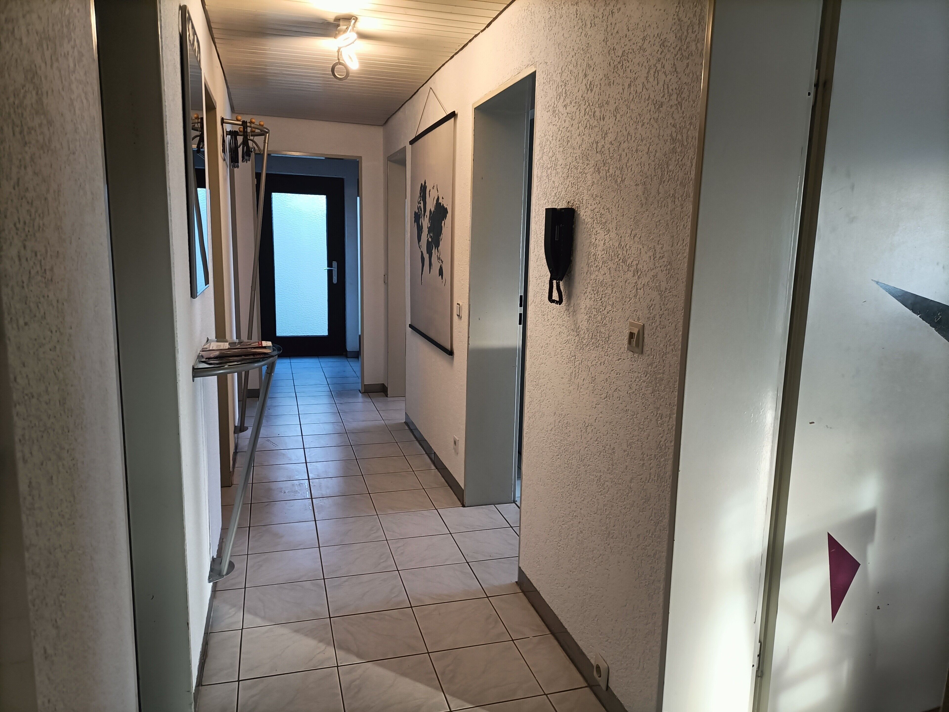 A hallway with tiled flooring, a ceiling fan, a door with a light switch, and a wall-mounted light fixture.
