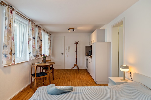 Ein kleines Zimmer mit Bett, Schreibtisch, Stuhl, Nachttisch, Lampe, Kleiderhaken und Kühlschrank.