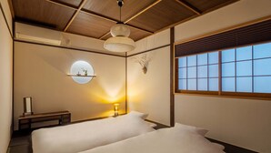 Ein Zimmer mit zwei Betten, einem Nachttisch, einer Lampe, einem an der Wand befestigten Regal und einem Fenster mit Jalousien.