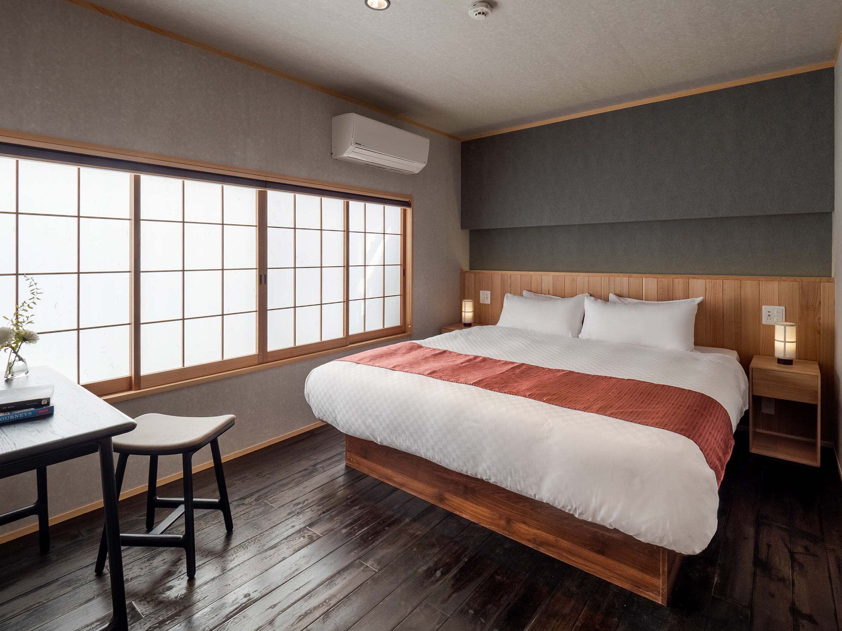 Hotellihuone, jossa on suuri ikkuna, puinen päätyosaan varustettu sänky, yöpöytä lampun kanssa ja pieni jakkara.
