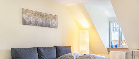 A modern loft bedroom with a gray sofa bed, a large window with a view, and a painting on the wall.