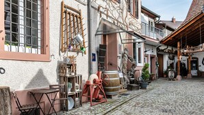 Een geplaveid straatje met gebouwen, een houten tafel, stoelen en diverse objecten zoals een vat en een houten ladder.
