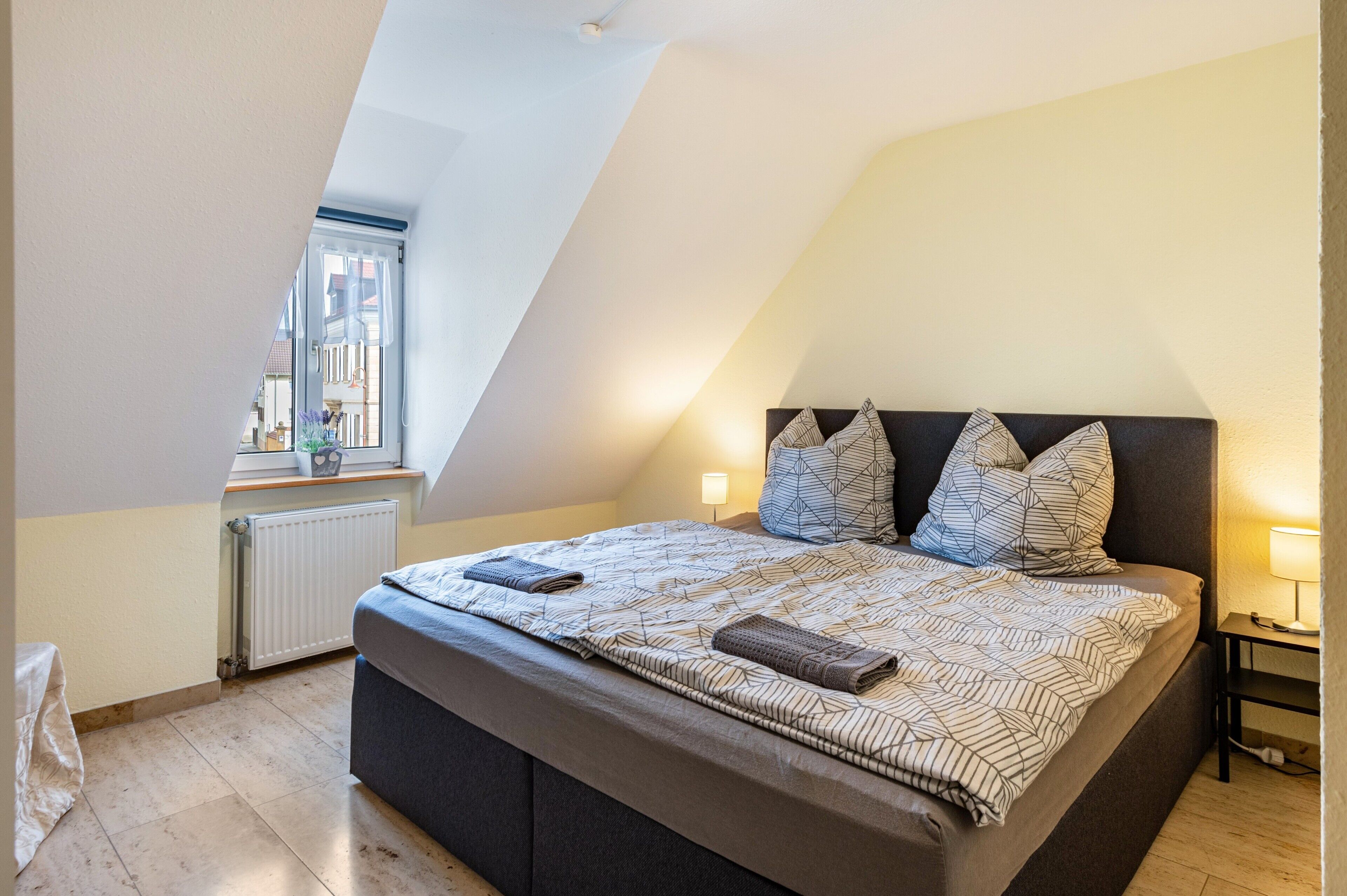 Een slaapkamer met een groot bed, een raam, een radiator en een nachtkastje met een lamp.