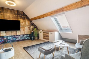 Una habitación tipo loft con techo inclinado, un televisor grande montado en la pared, una pequeña cocina americana y una zona de estar con mesa y sillas.