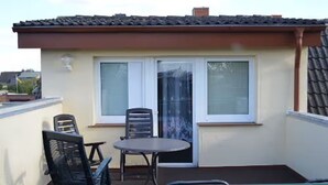 Ein kleiner Balkon mit Tisch und zwei Stühlen, ein Fenster mit Rollläden und ein Ziegeldach.
