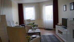 Ein Wohnzimmer mit Sofa, Sessel, Couchtisch, TV-Stand und einem großen Fenster mit Vorhängen.