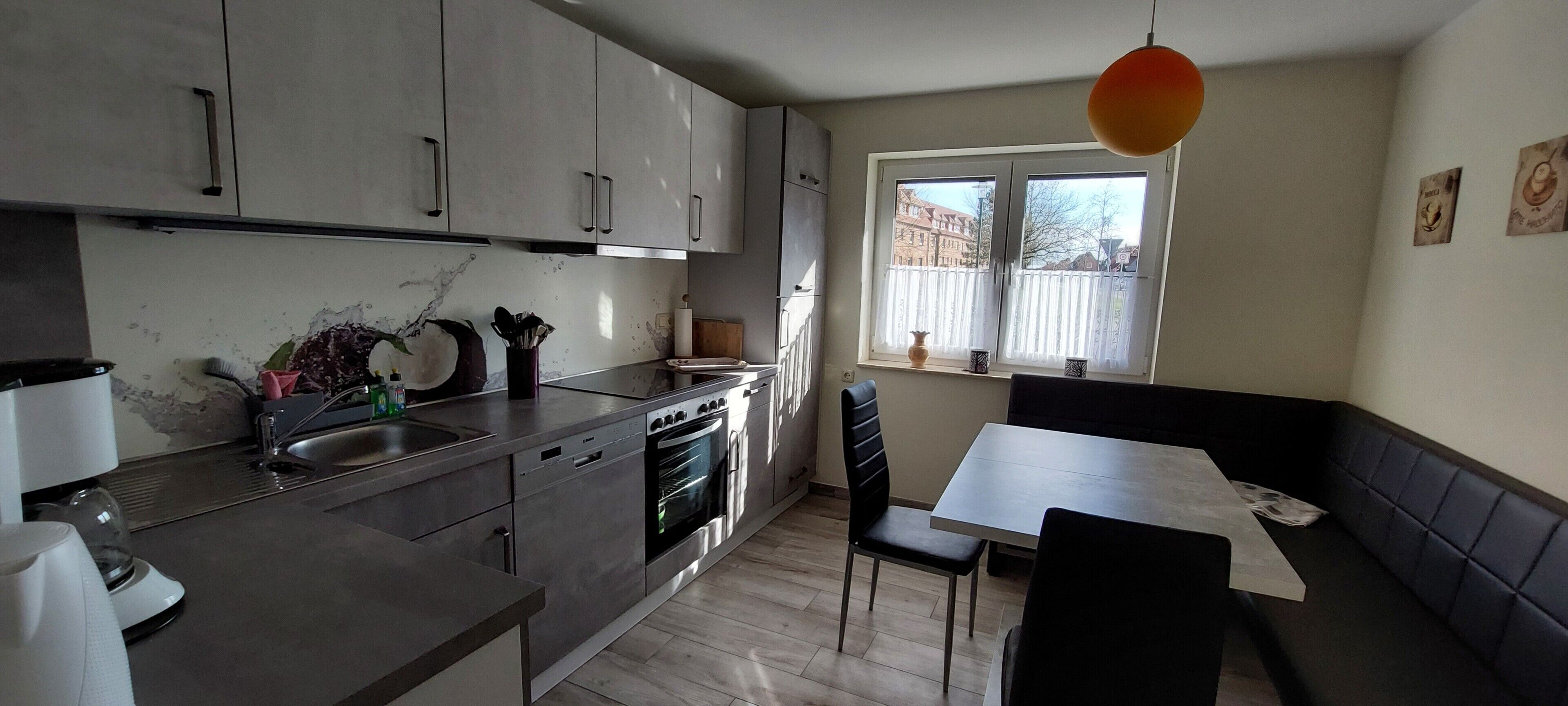 Une cuisine moderne équipée d’appareils électroménagers en acier inoxydable, un coin repas avec une table et des chaises, et une fenêtre donnant sur des immeubles.