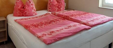 A bedroom with a white bed, pink floral bedding, a wooden headboard, a nightstand, and a window with natural light.