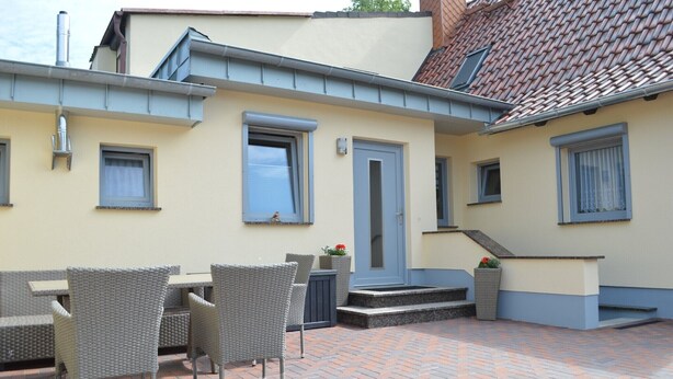 Een gebouw met een pannendak, een deur, ramen en een patio met stoelen en een tafel.