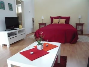 Une chambre à coucher avec un lit, un téléviseur posé sur un meuble, une petite table avec une plante et un tapis rouge.
