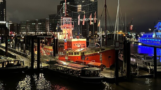 Un port de plaisance au bord de l’eau, avec un bateau rouge amarré, un bateau bleu en arrière-plan et un paysage urbain avec des bâtiments.