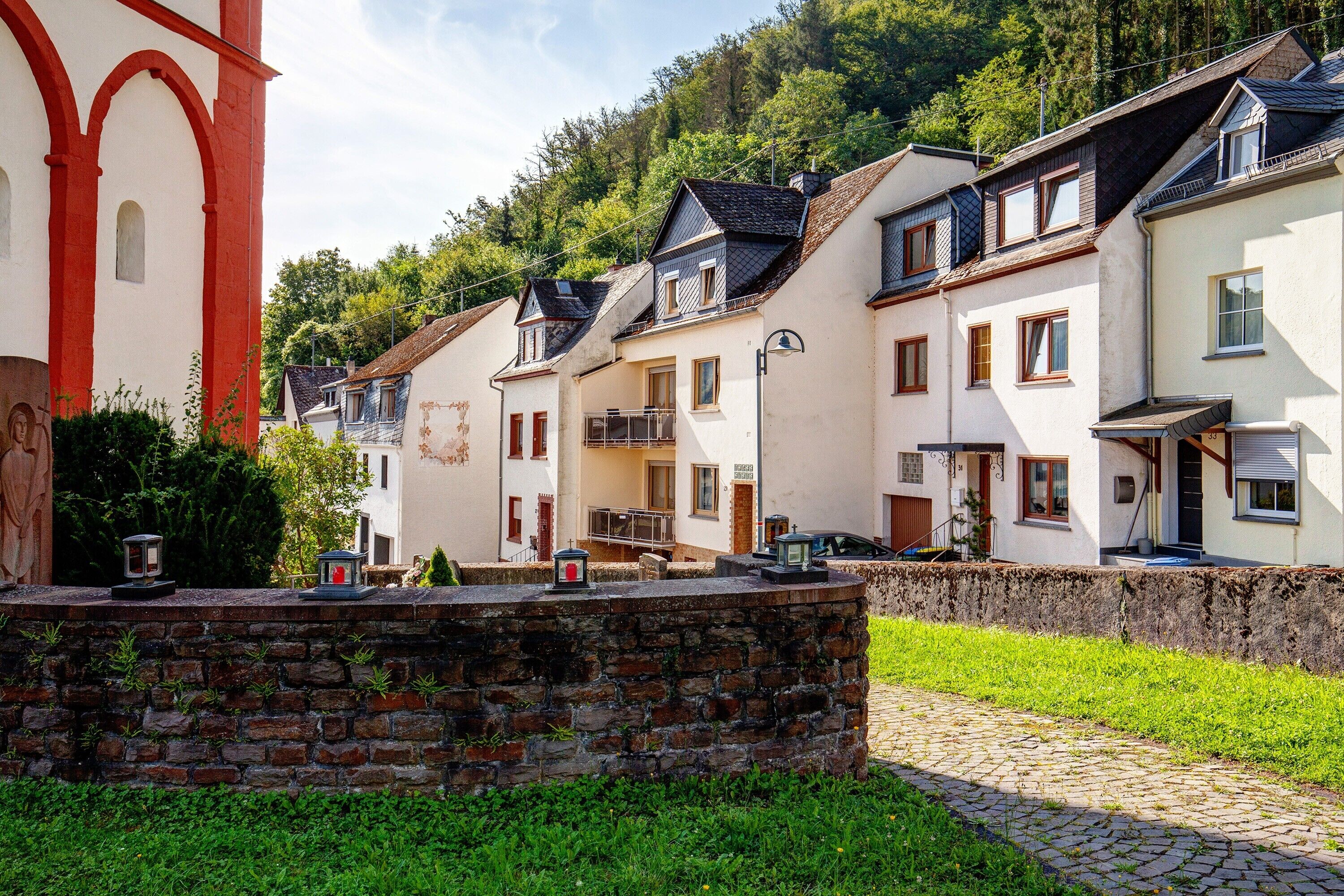 A row of traditional European-style buildings with a stone wall in the foreground, a cobblestone path, and a green lawn.