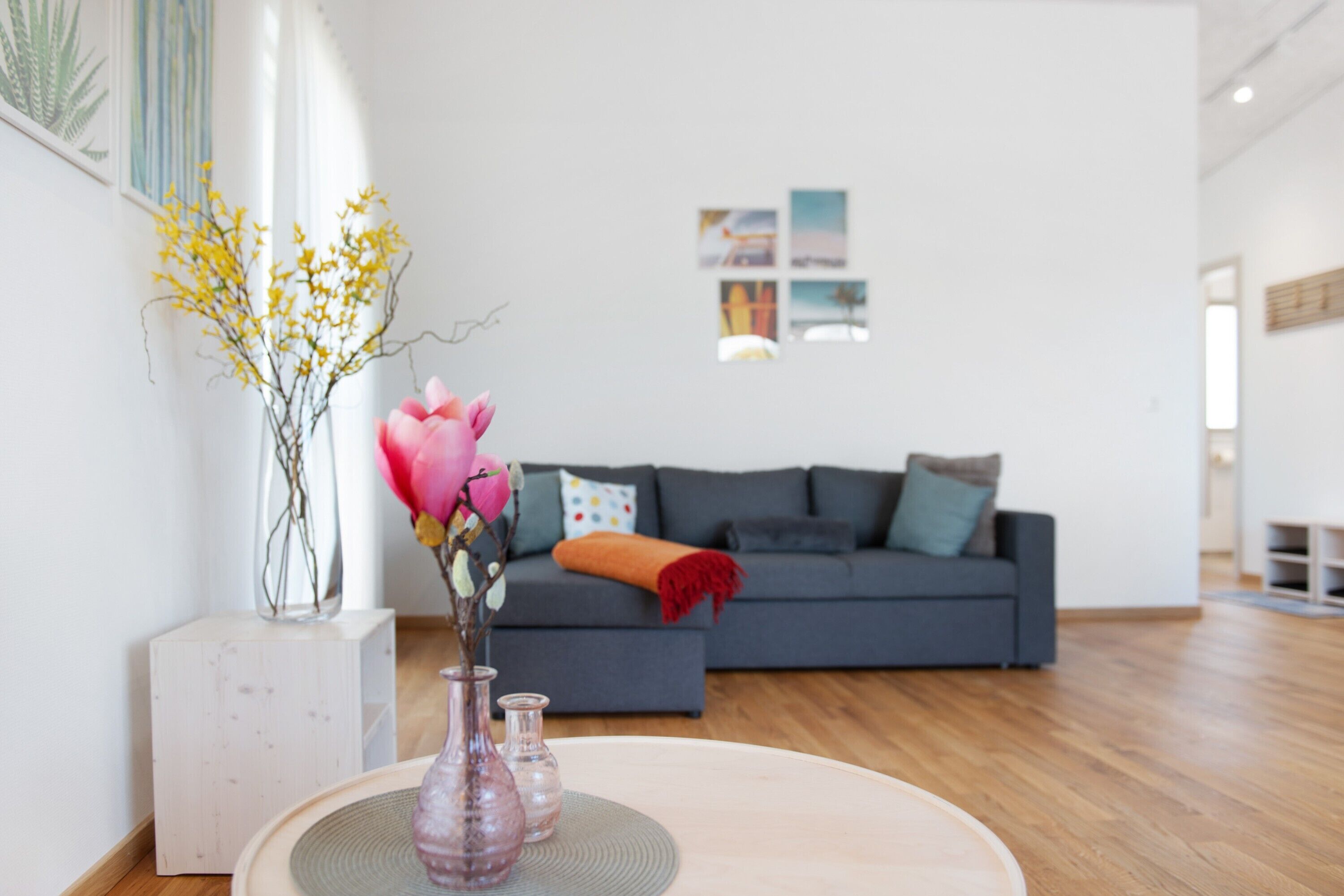 Un soggiorno moderno con un divano grigio, un tavolo rotondo in legno con un vaso di fiori e un vaso bianco con fiori gialli.