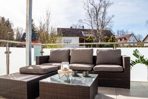 Un balcon avec un canapé en osier, une table basse avec un plateau en verre et une rambarde en métal.