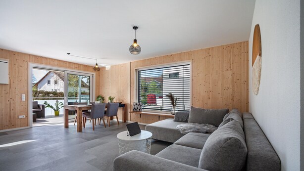 A modern living room with a sofa, dining table, and chairs, a small round table with a laptop, and a large window with blinds. - Holiday Apartment 'Wohnen an der Ach' with Private Garden, Balcony and Wi-Fi (Bad Wurzach)