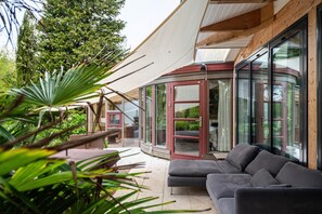 Een moderne buitenruimte met een gebogen glazen constructie, een grijze bank en een houten terras.