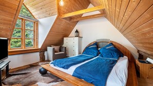 Quarto com teto inclinado, cama com roupa de cama azul, cómoda, televisão e janela com vista para área verde.