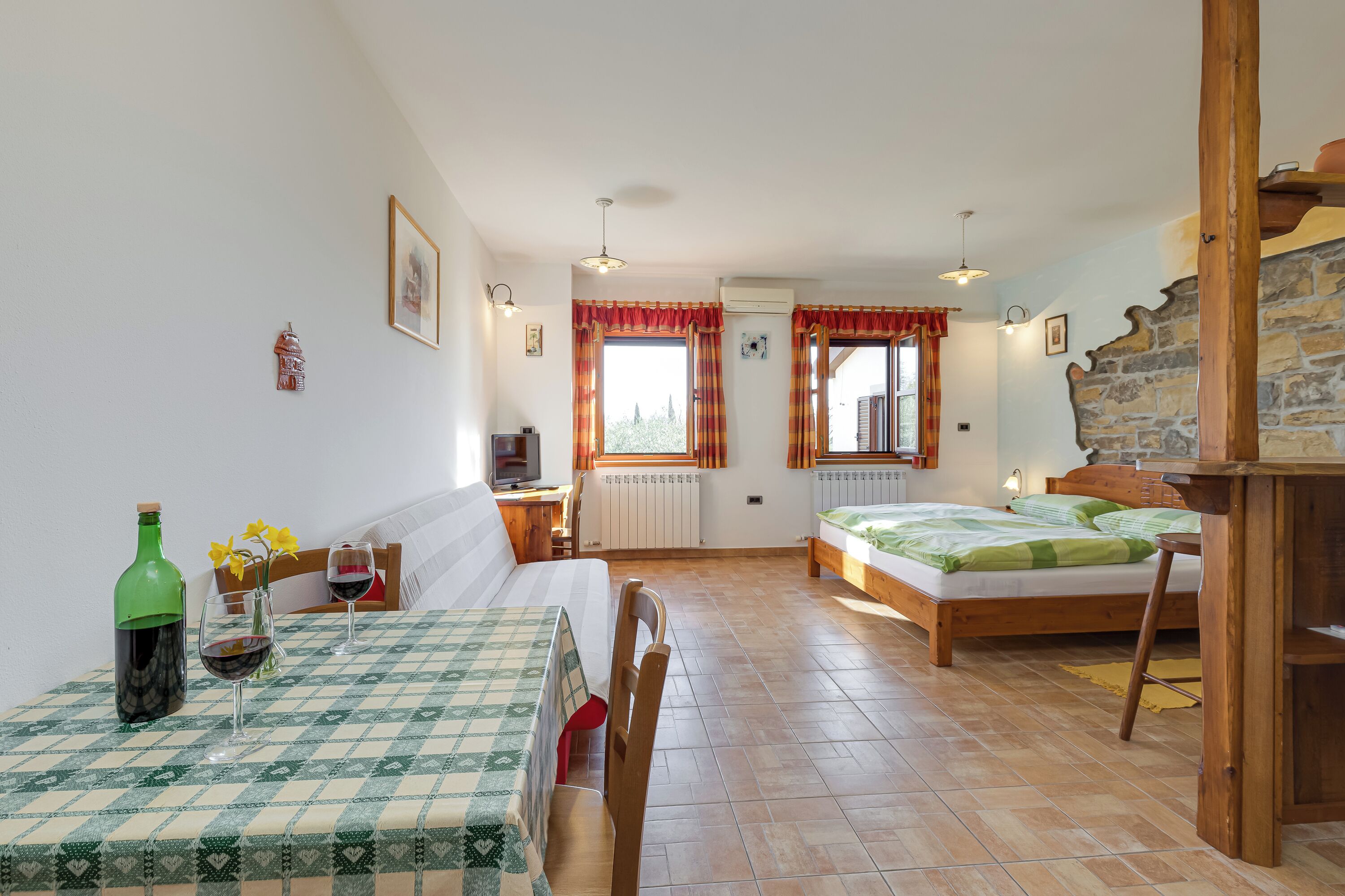 A room with a bed, a table with a checkered tablecloth, a chair, a radiator, and a window with curtains.