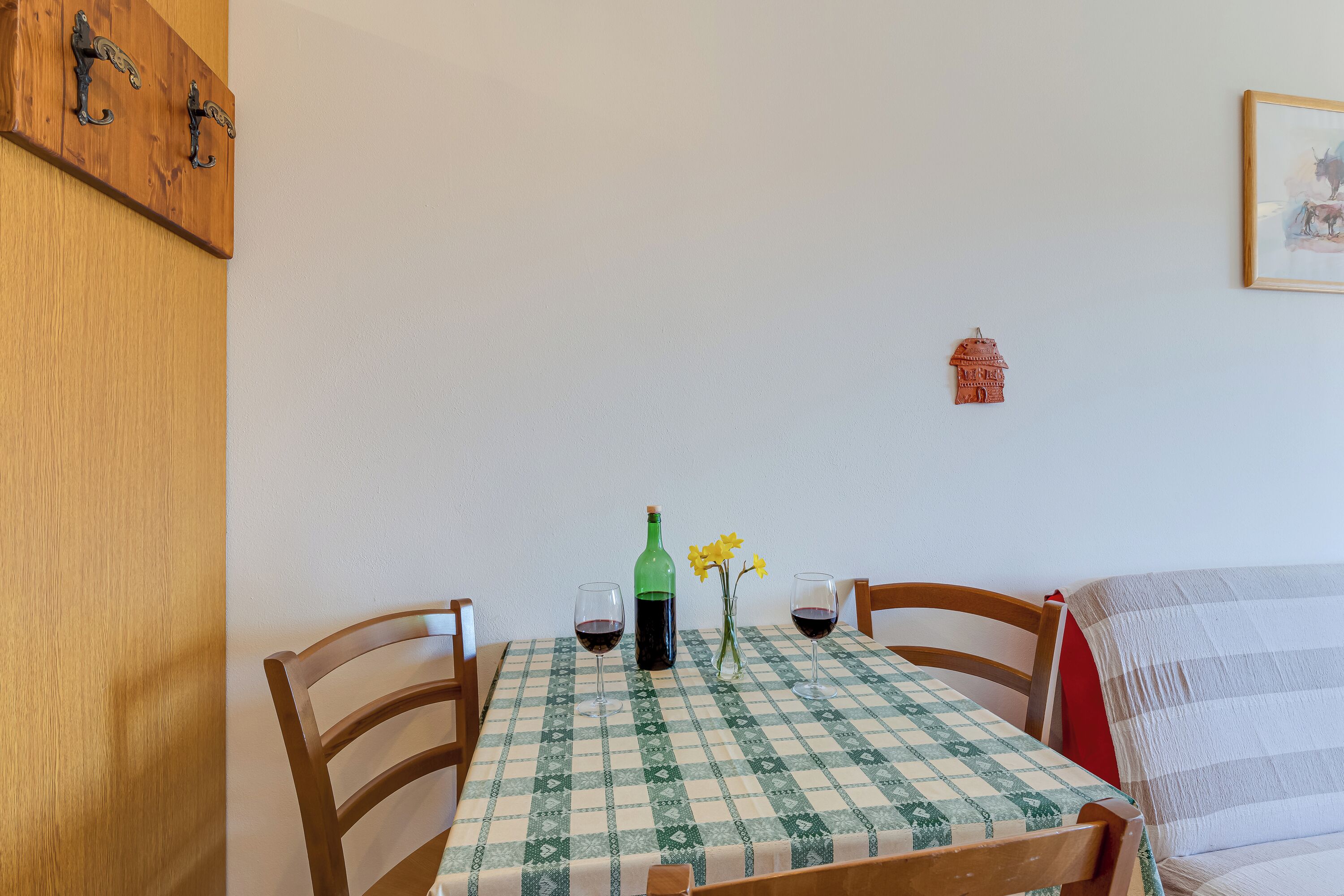 A dining table with a checkered tablecloth, two chairs, a bottle of wine, and a vase with flowers.