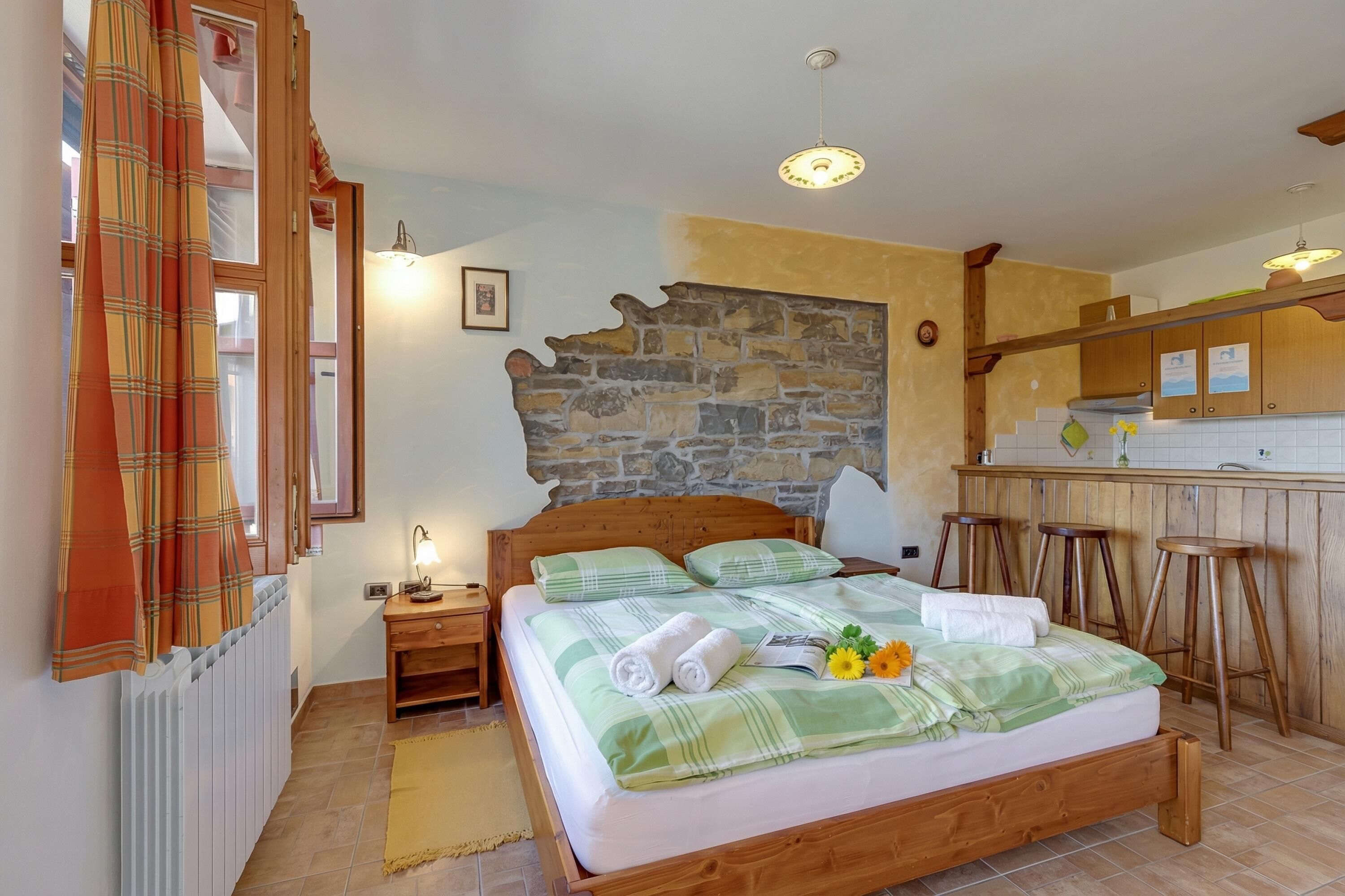 A bedroom with a wooden bed, a stone wall, a kitchen area with wooden stools, and a radiator.