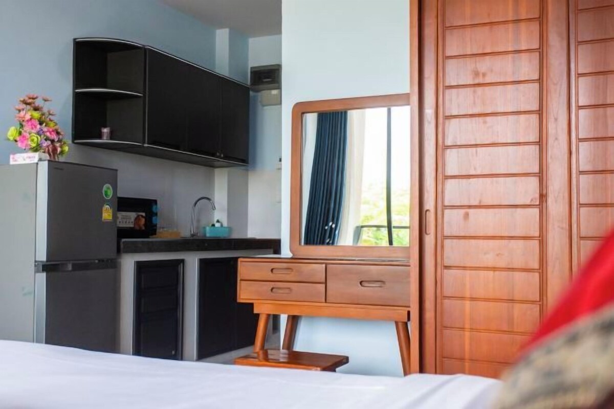 A compact hotel room with a bed, a wooden dresser with a mirror, a small kitchenette with a refrigerator, and a wooden wardrobe.