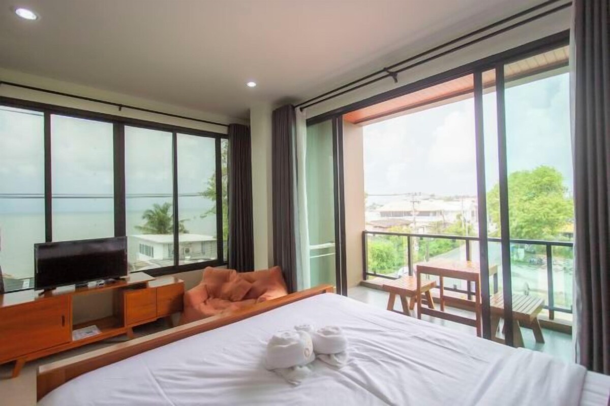 A modern hotel room with a large bed, a TV, and a balcony with a view of the sea.