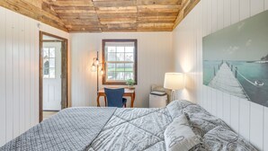 Et soveværelse med træloft, seng med mønstret sengetøj, et skrivebord med stol, et vindue og et billede af en bro.