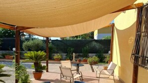 An outdoor patio with a round table and chairs, surrounded by potted plants and a shaded area.