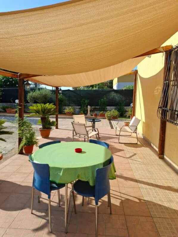 An outdoor patio with a round table and chairs, surrounded by potted plants and a shaded area.