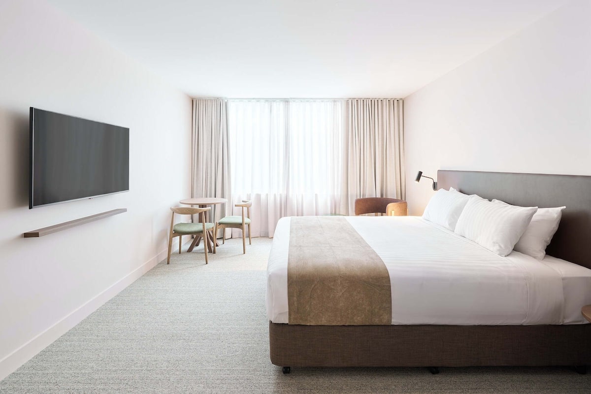 A hotel room with a large bed, a flat-screen TV mounted on the wall, a small table with two chairs, and a window with curtains.